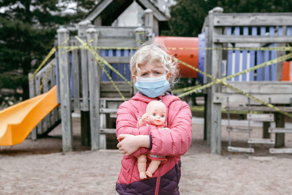 Child wearing medical mask in front of closed playground representing school closures and learning loss from COVID-19 pandemic.