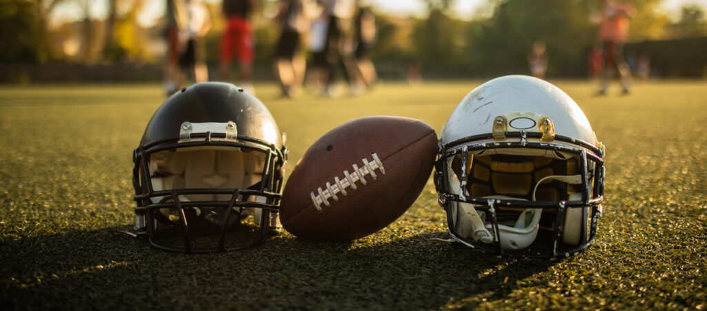 Details Of American Football Equipment: Football Helmet And Ball Beside It, On Sunny Stadium