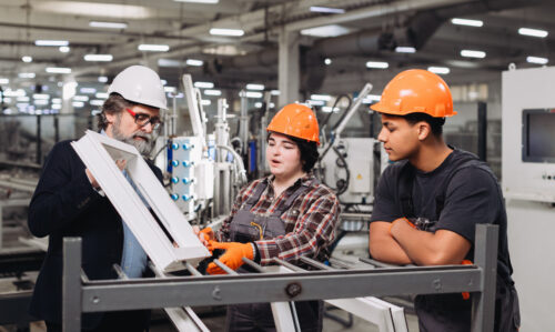 Mentor and young workers inspecting window frame, discussing production process and quality control in a manufacturing facility apprenticeship week