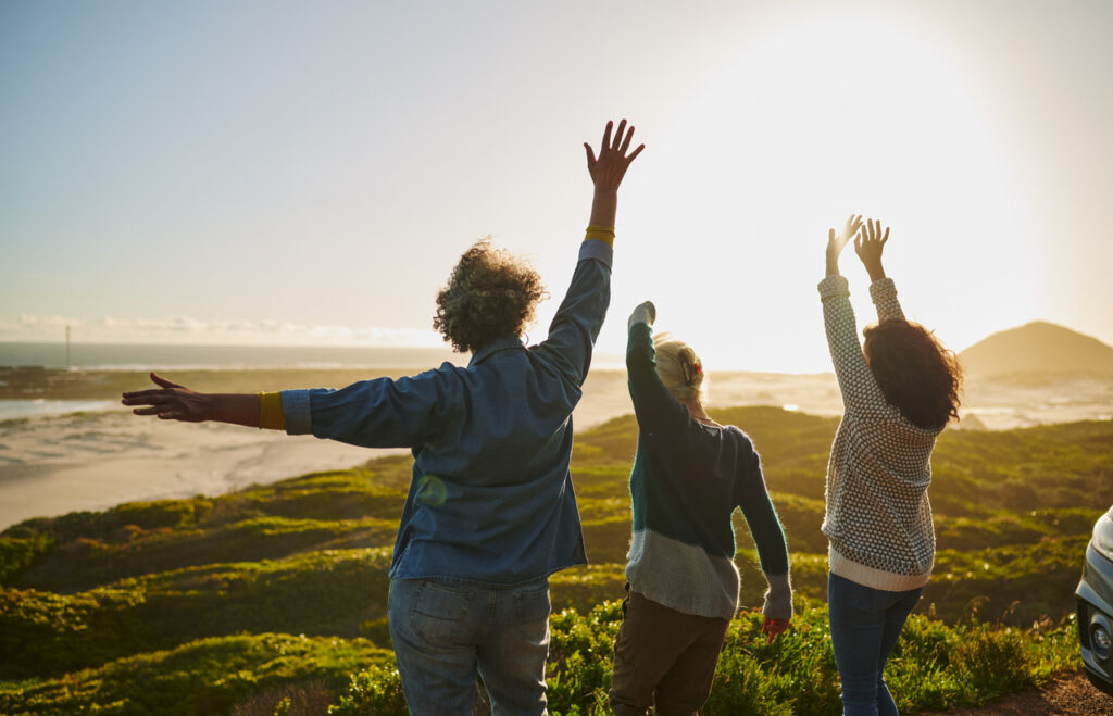 Rear view of a group of mature female friends cheering with their arms raised by their car parked on a road running along a scenic coastline